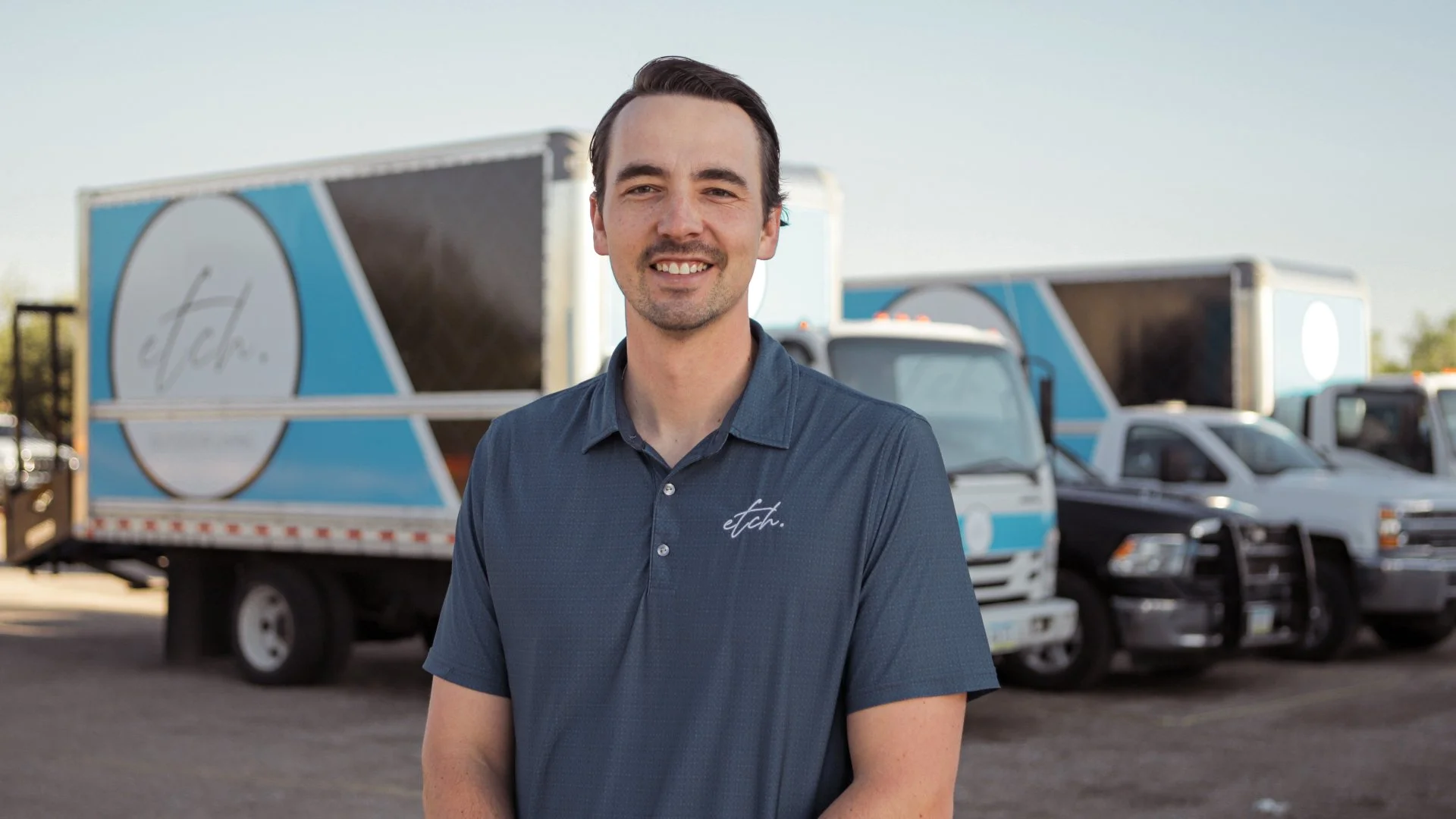 Individual standing in front of a fleet of branded delivery trucks, smiling, and wearing a company polo shirt.